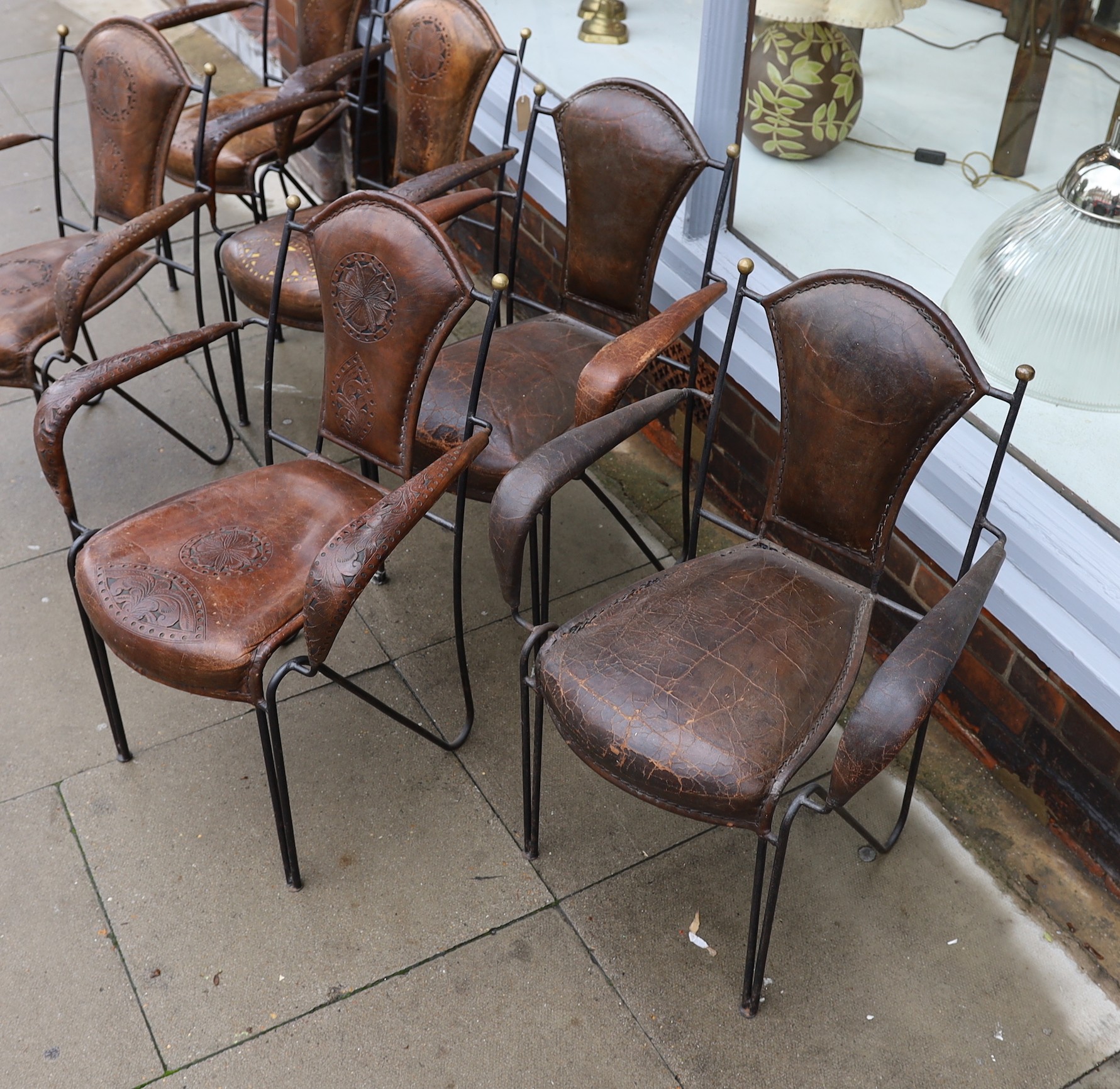 A set of six French wrought iron and brown leather elbow chairs, with brass ball finials, four with tooled and pierced decoration the others plain, height 84cm. width 61cm